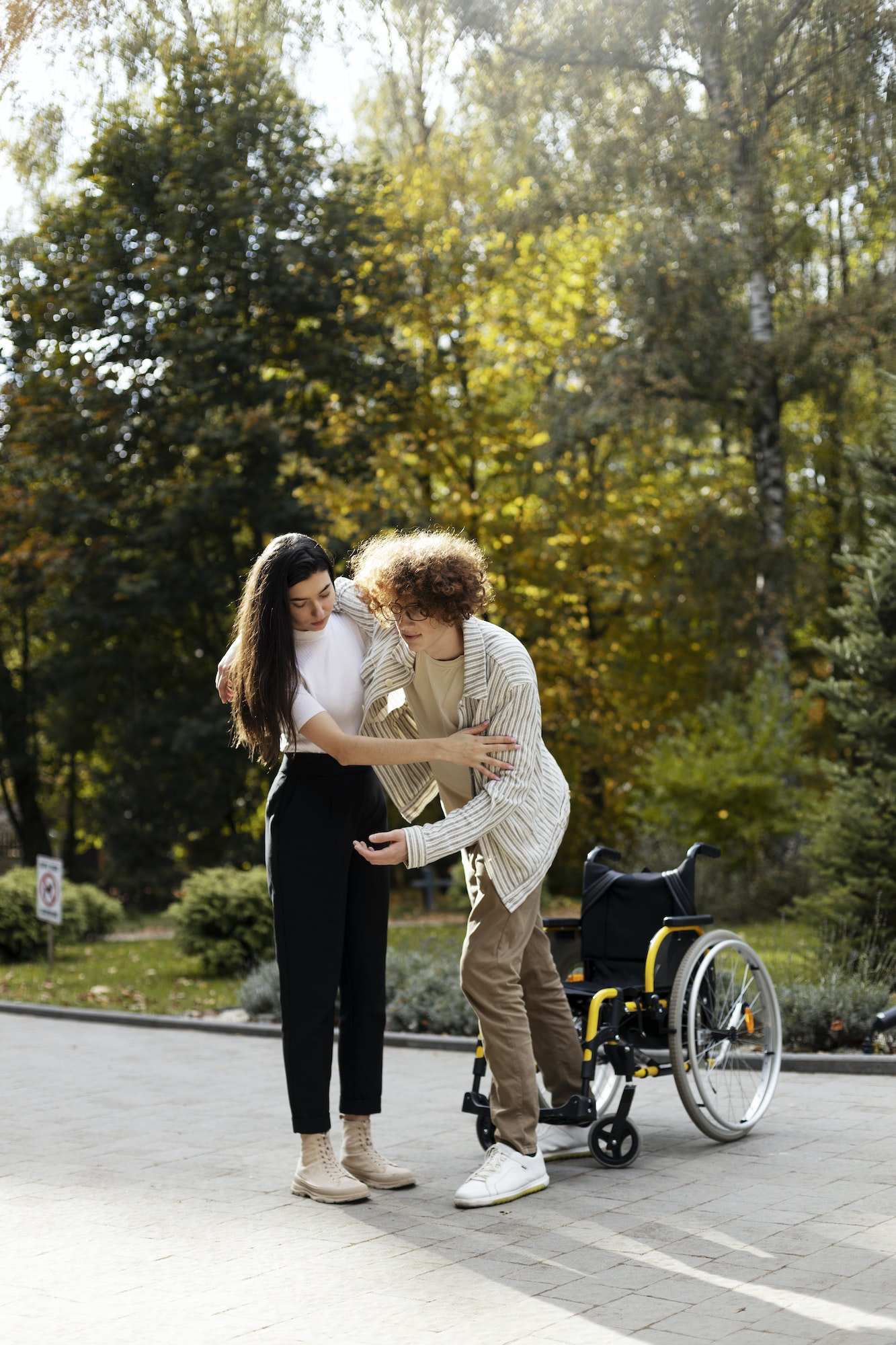 Attractive, young girl helps a young man to get up from a wheelchair. Young couple outdoors.